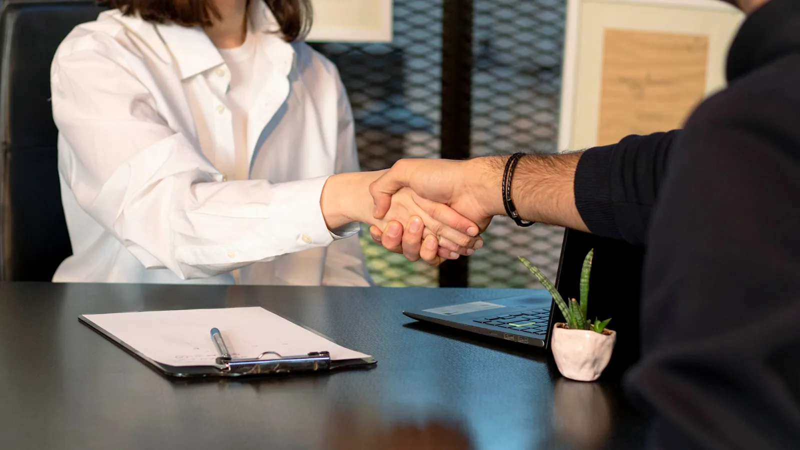 Two people shaking hands in front of an open laptop in an office